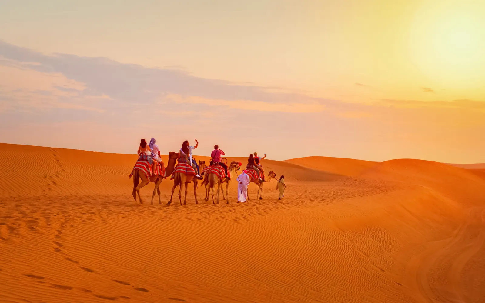 Camels and riders crossing orange sand dunes at sunset