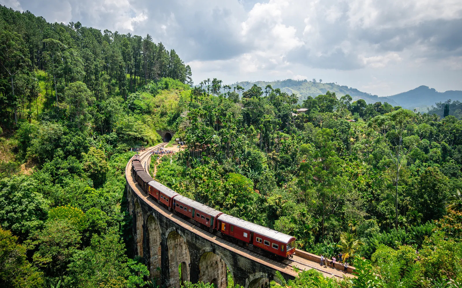 A red train crossing a high stone viaduct surrounded by jungle
